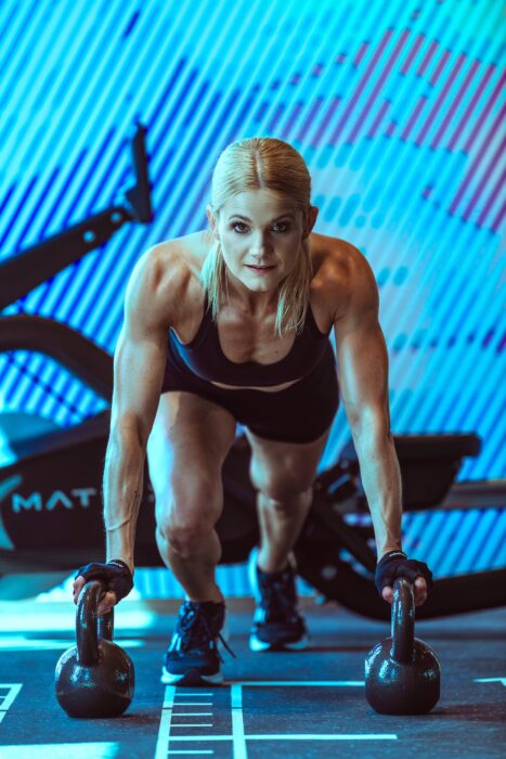 Athletische Frau trainiert mit zwei Kettlebells in einem Fitnessstudio.